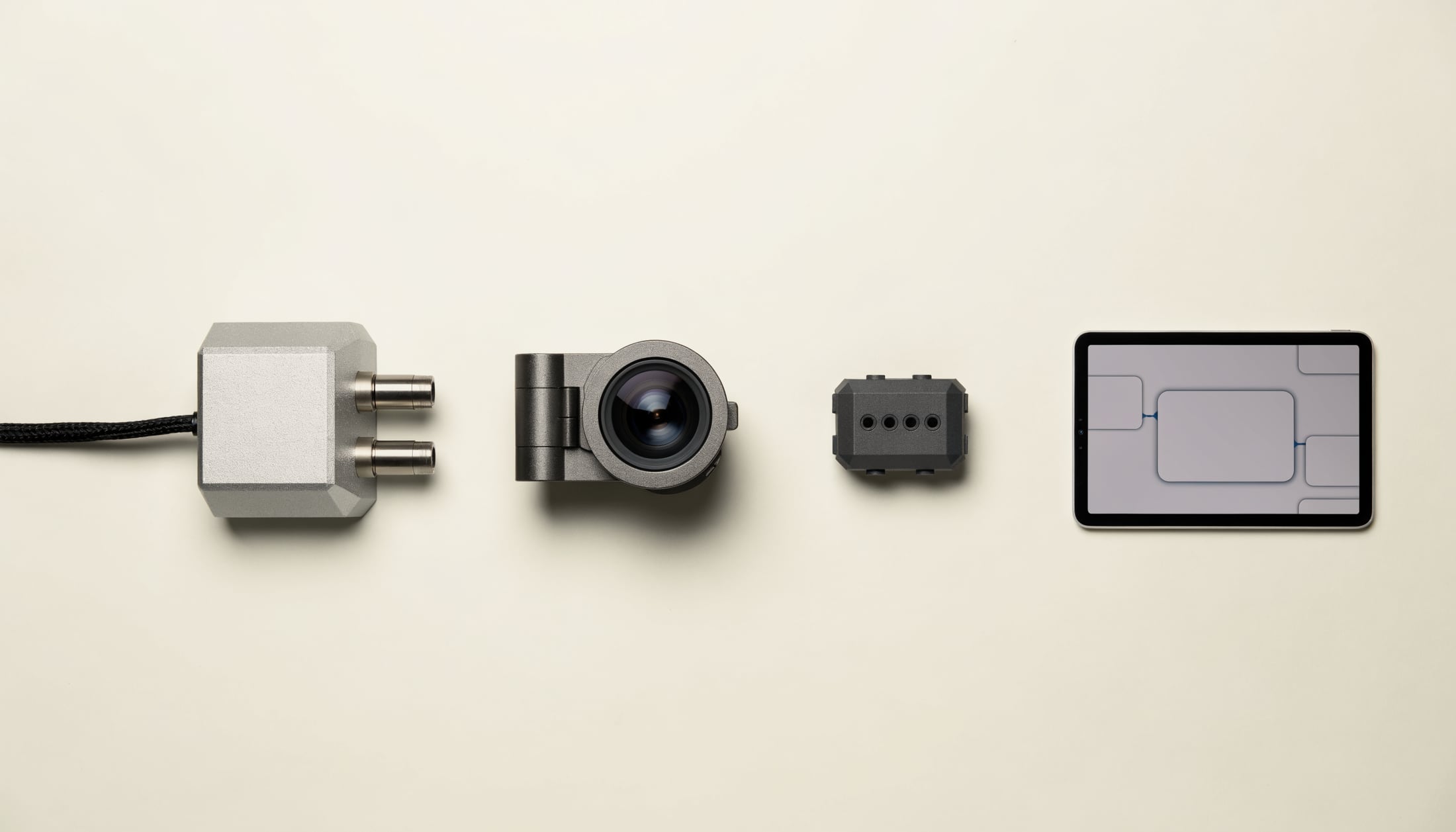 Product photograph: four Olytoma products arranged in a row on a pale warm surface — an industrial sensor node with probe fittings and a braided cable, a machined clip-on camera module with a large glass lens, a ruggedized sensor head, and a tablet showing an interface layout.