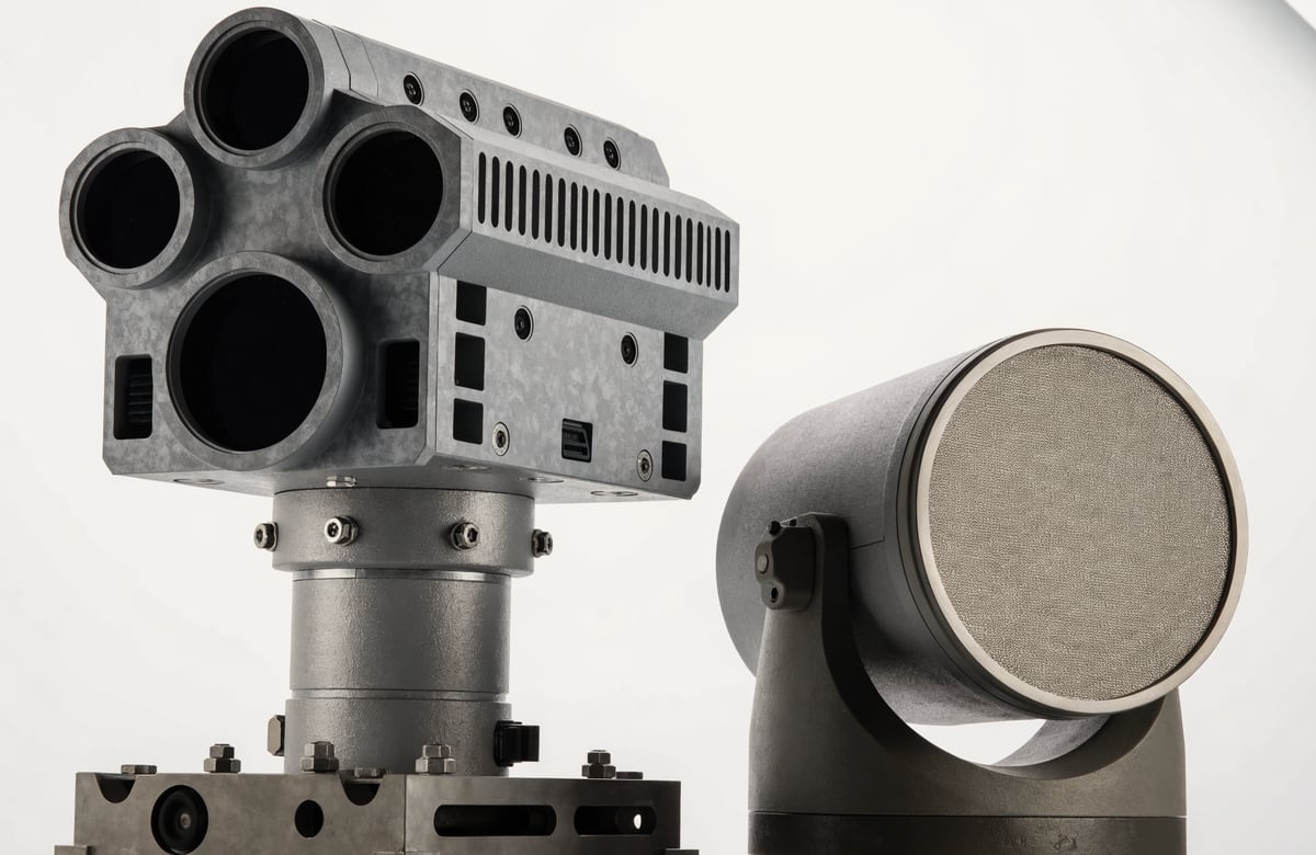 Product photograph: a ruggedized multi-aperture passive sensor head mounted on a short mast baseplate, beside a cylindrical effector unit on a low pan-tilt cradle with a fine mesh emitter face.